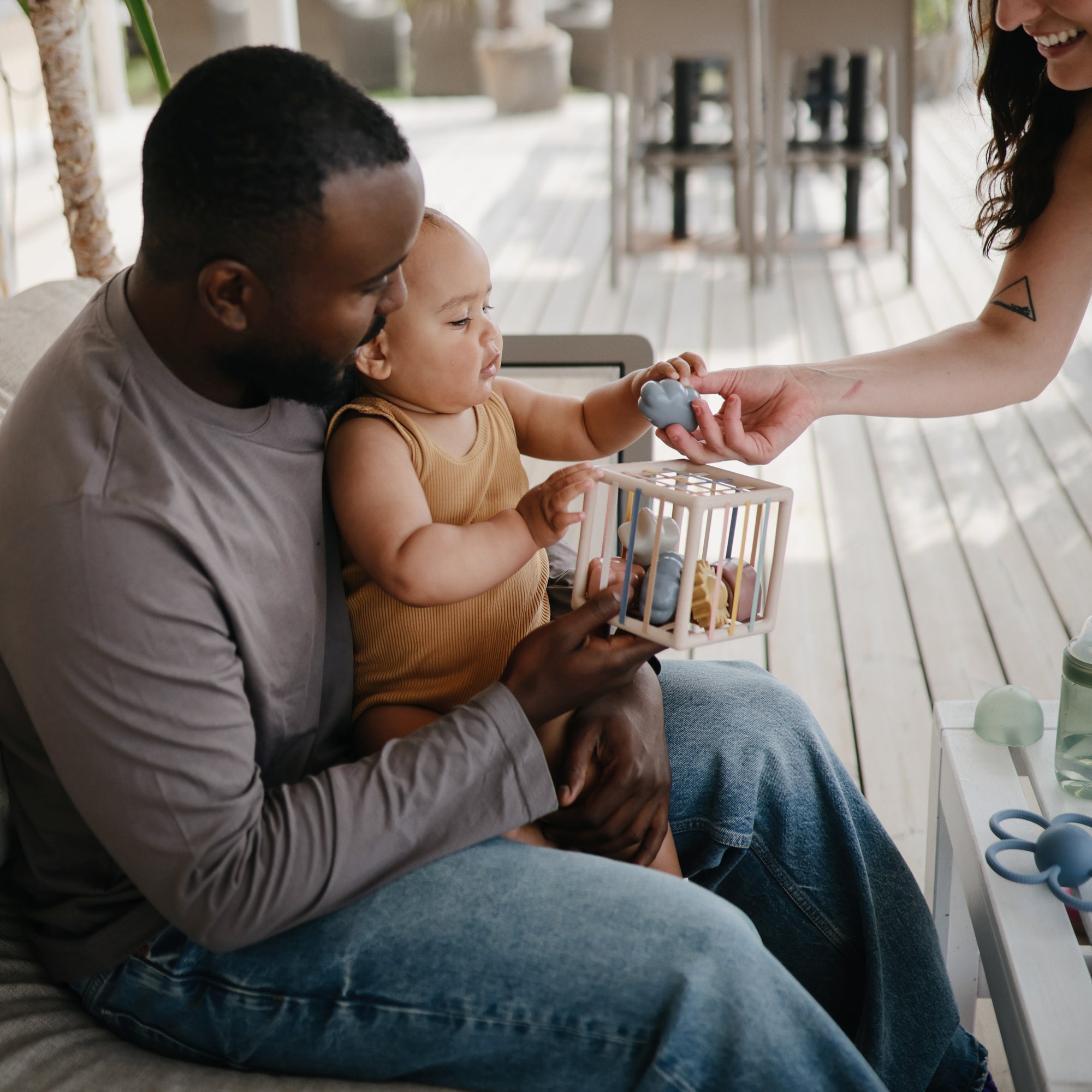 A man holds a baby on his lap while the baby reaches for a toy being handed by a woman. They are sitting together on a porch, and the baby is wearing a brown outfit, playing with a wooden toy.