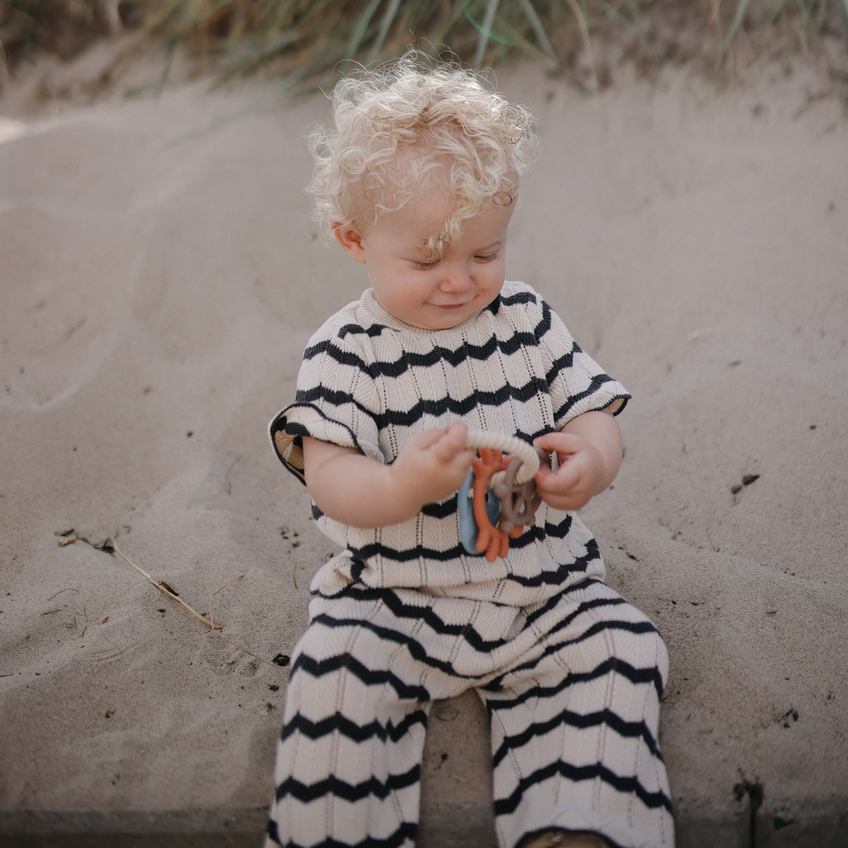 A toddler in a striped outfit sits on sand holding a mushie Teething Ring.