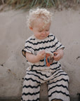 A toddler in a striped outfit sits on sand holding a mushie Teething Ring.