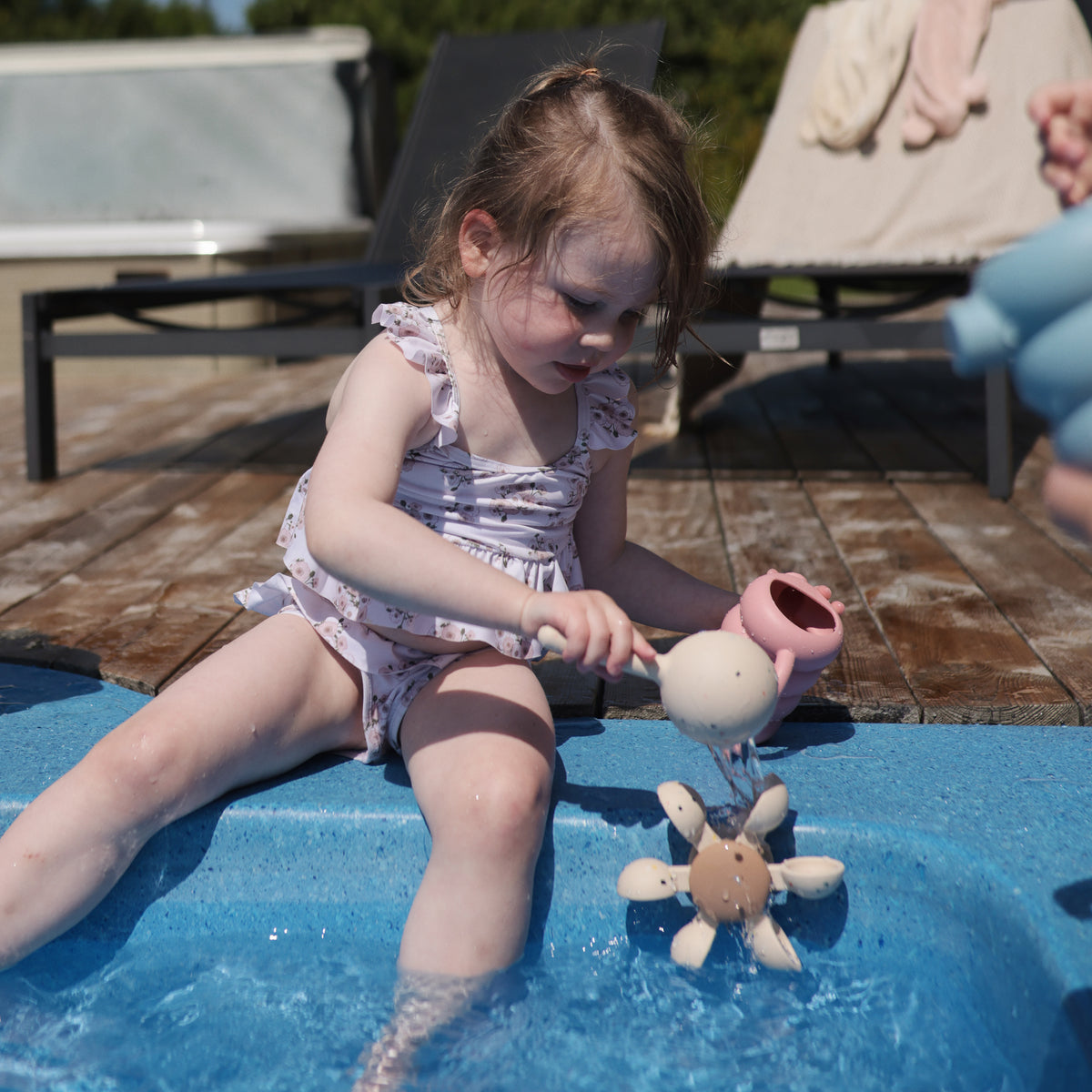 Child by pool pours water on mushie Water Wheel Bath Toy in swimsuit.