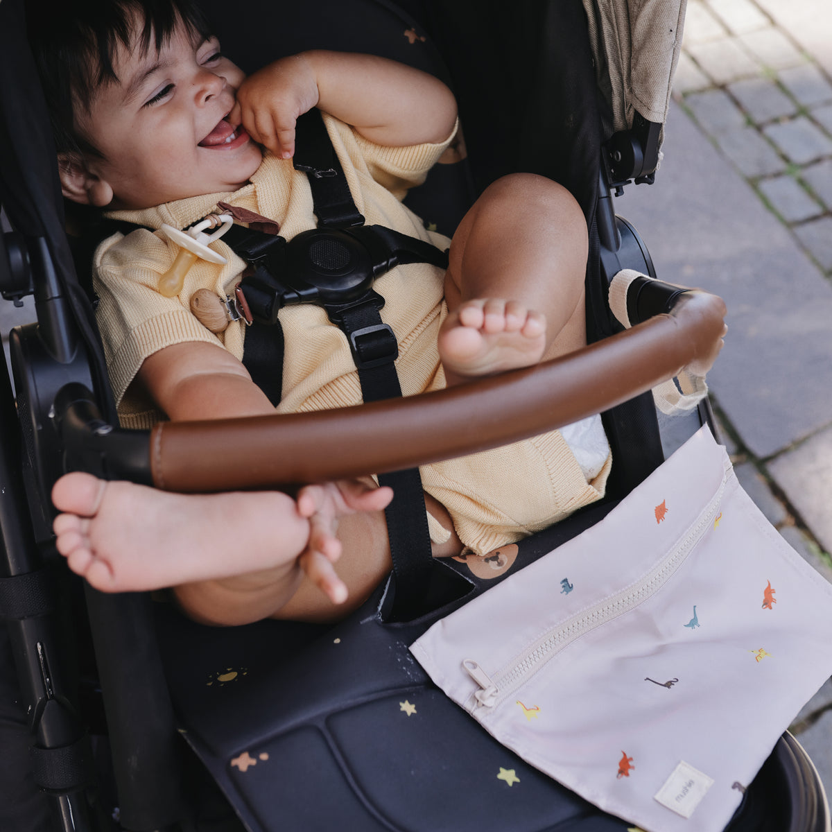 A happy baby in a stroller with a mushie Water Resistant Wet Bag featuring dinosaurs and stars.