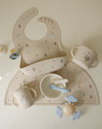 Mushie Silicone Baby Bib on a matching placemat with cups, bowl, utensils & teether.