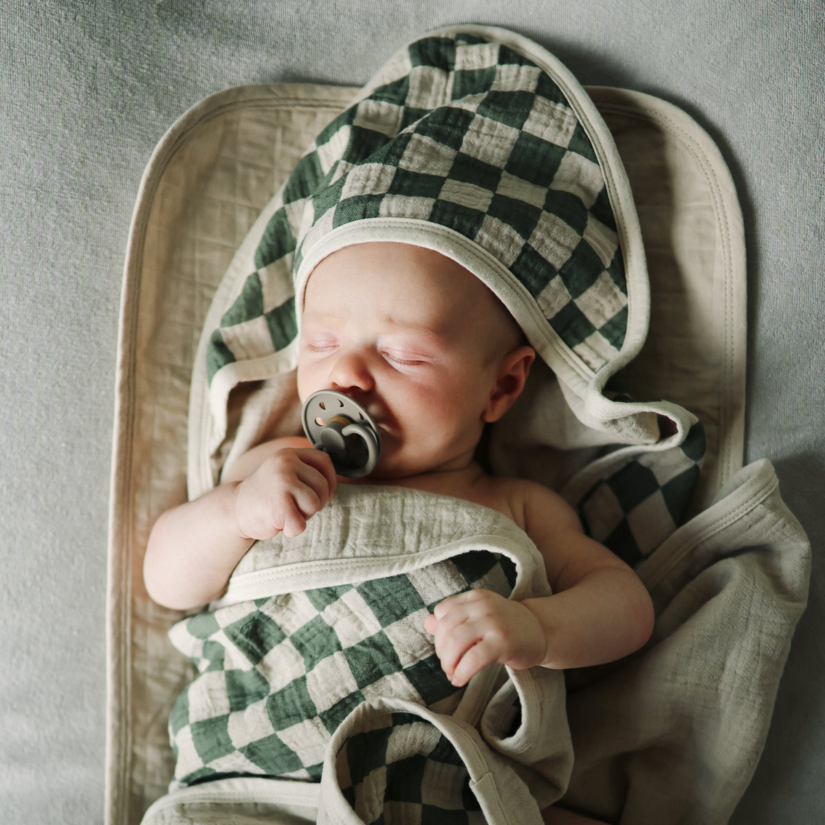 A baby with a pacifier sleeps on Mushie Changing Pad Liners in a checkered hooded towel.