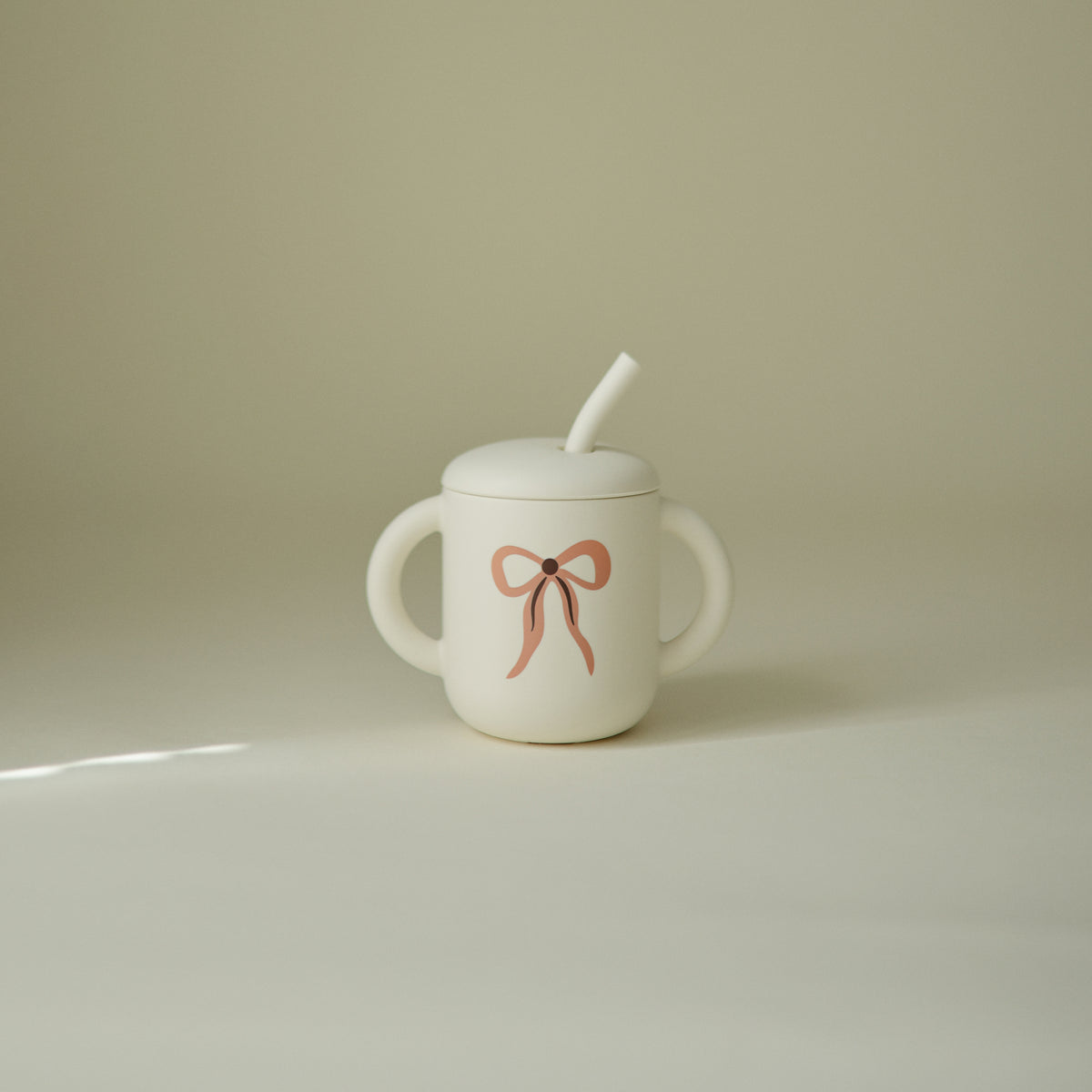 Mushie Silicone Training Cup + Straw: white, two handles, lid, pink bow design.