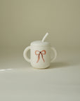Mushie Silicone Training Cup + Straw: white, two handles, lid, pink bow design.