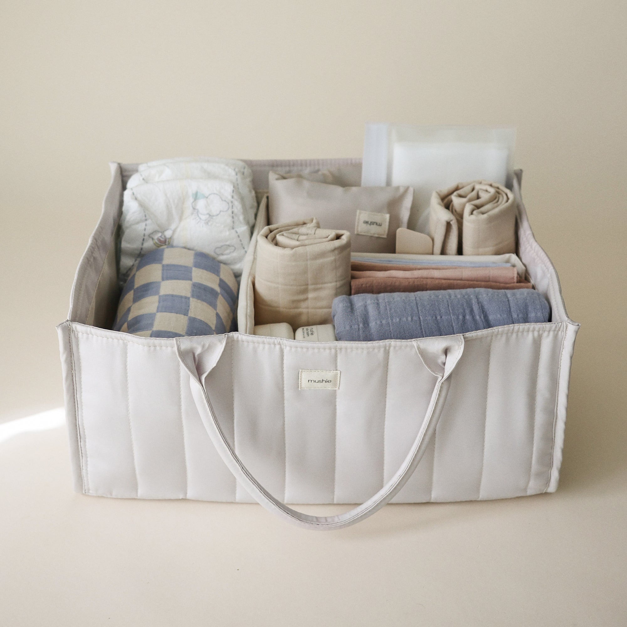 Lifestyle image of the Ivory Diaper Caddy with folded clothes and baby nursery items on a beige background. 