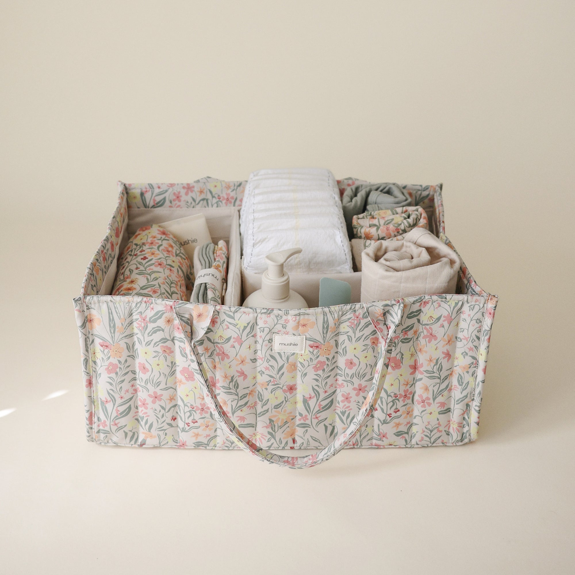 Lifestyle image of the Pastel Blooms Diaper Caddy with folded clothes and baby nursery items on a beige background. 