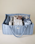 Lifestyle image of the Tradewinds Diaper Caddy with folded clothes and baby nursery items on a beige background. 