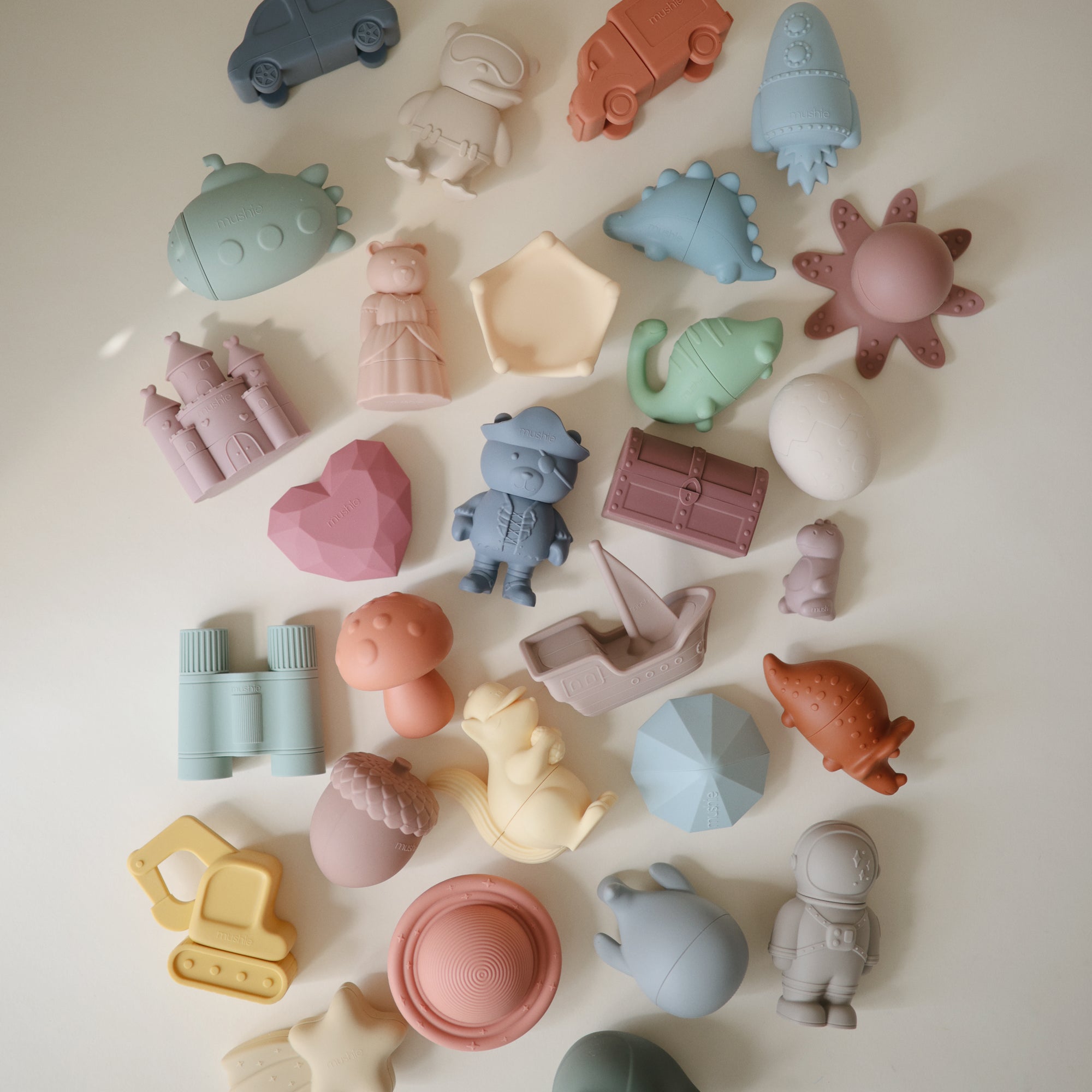 Lifestyle image of a collection of colorful silicone bath play set toys on a light background. The mix includes playful shapes from the Sealife, Pirate, Princess, Vehicles, Space and Dinosaur sets. 