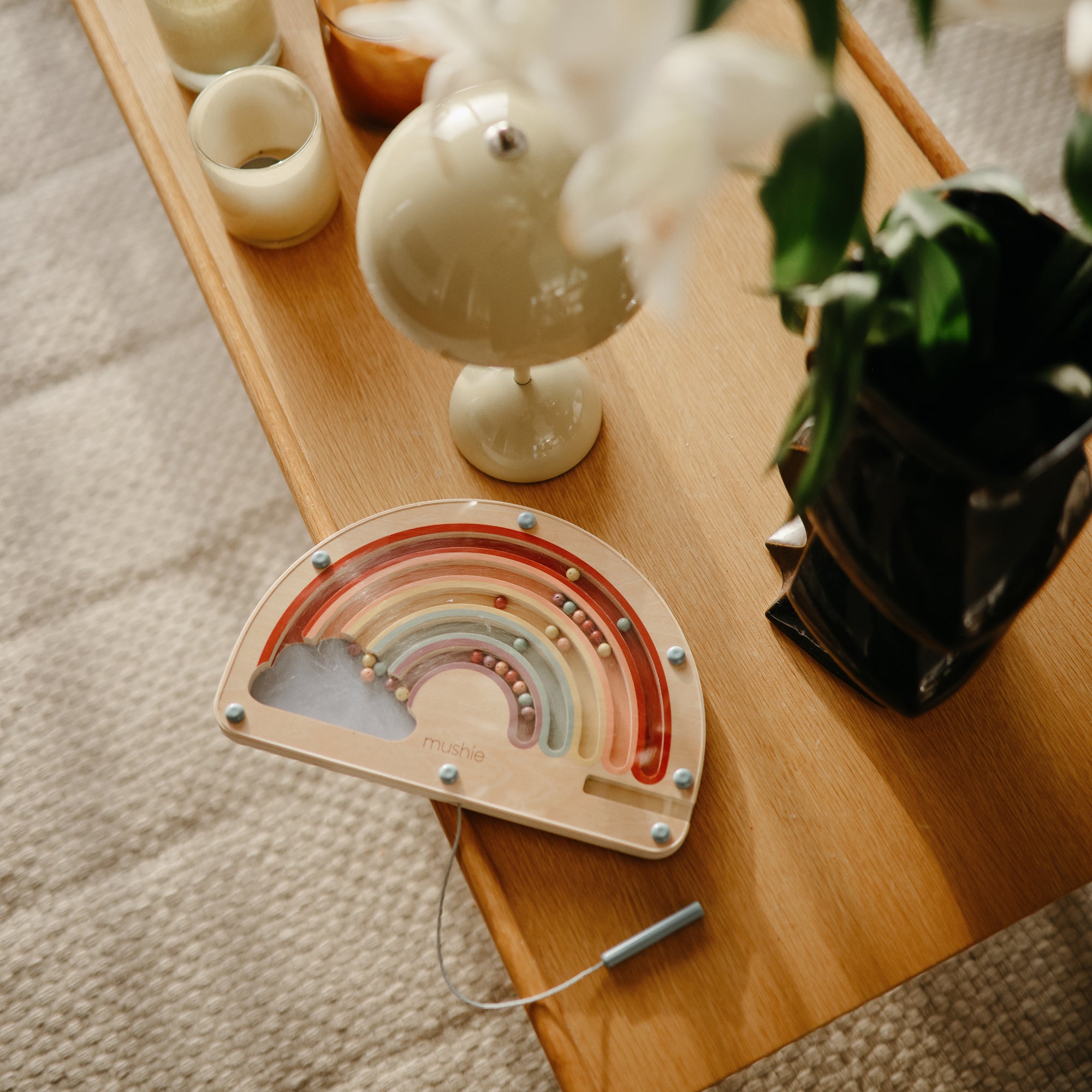 The mushie Wooden Magnetic Rainbow Maze rests on a light wood table by candles, a lamp, and a potted plant atop a soft rug—ideal for screen-free fun and developing fine motor skills.