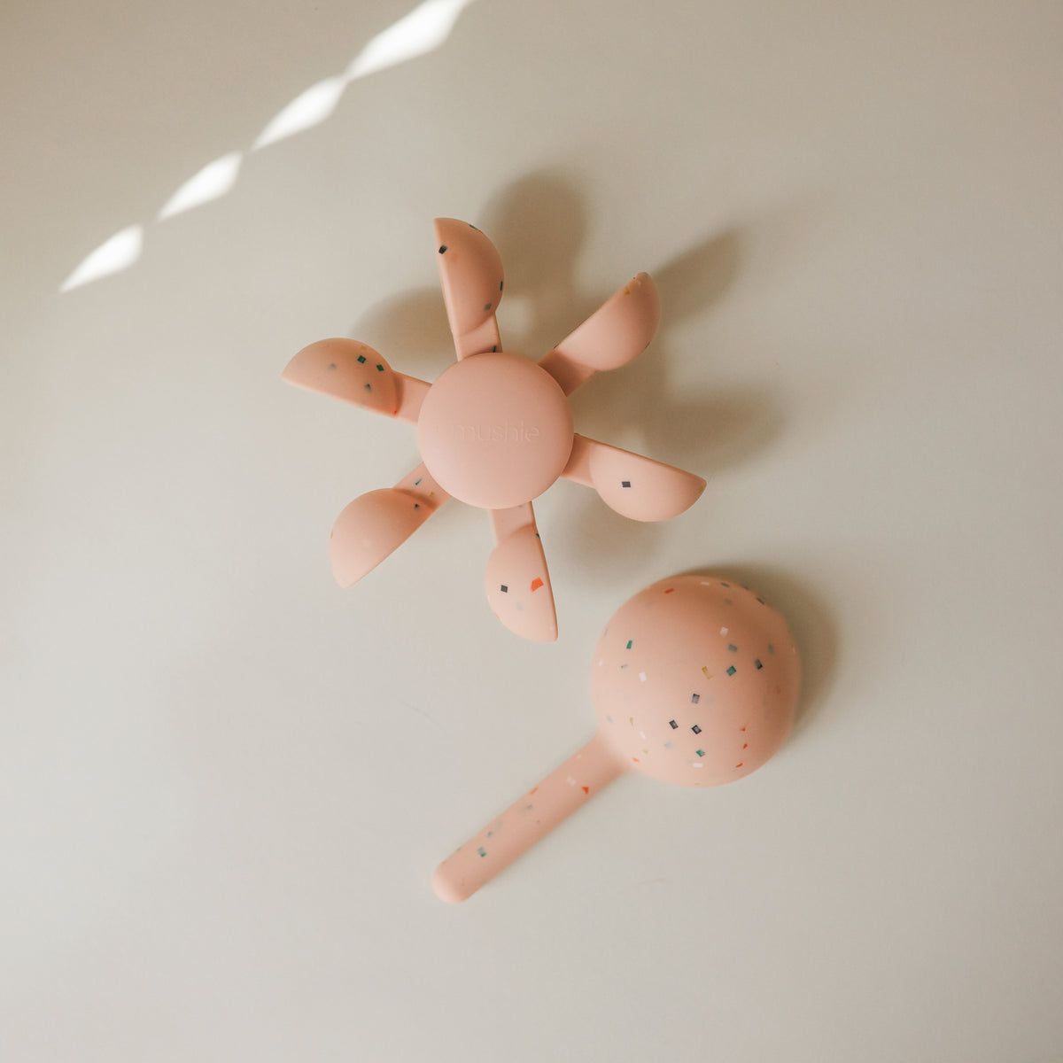 Two mushie Water Wheel Bath Toys, pink round and flower, speckled, on a beige surface.