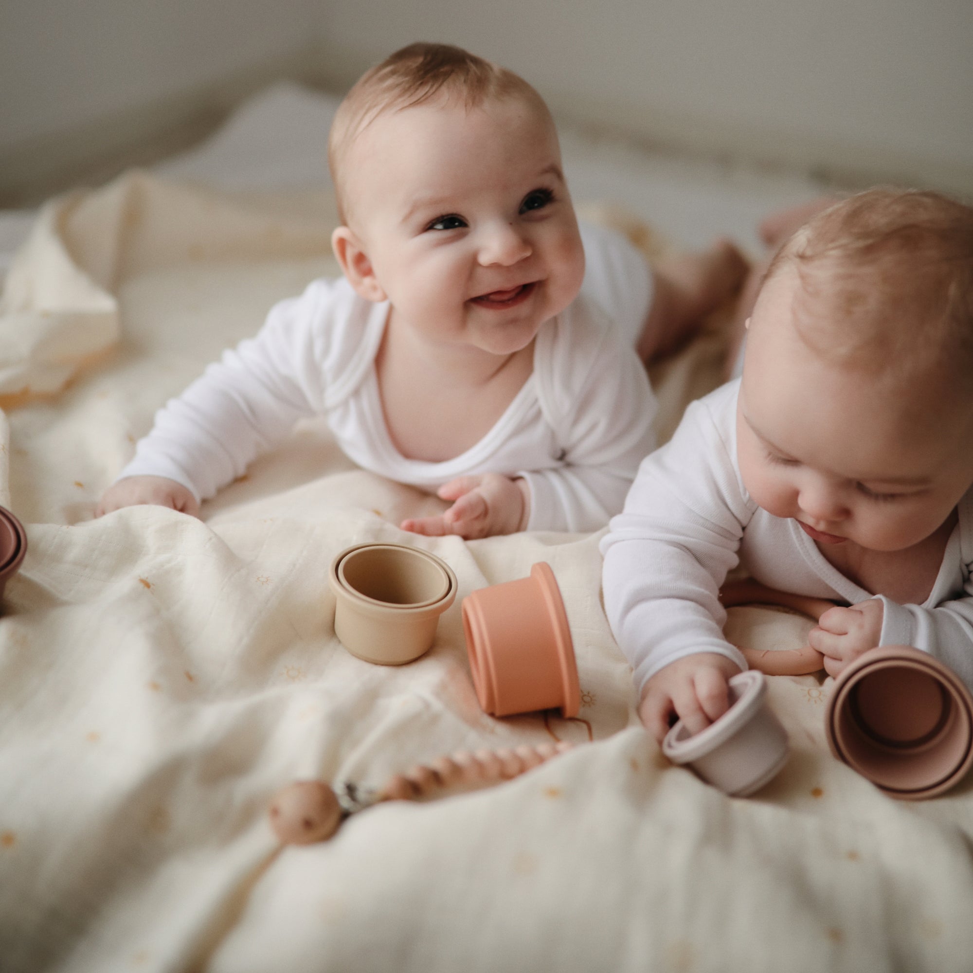 Two babies play with stacking cups on a mushie Organic Cotton Muslin Swaddle Blanket.