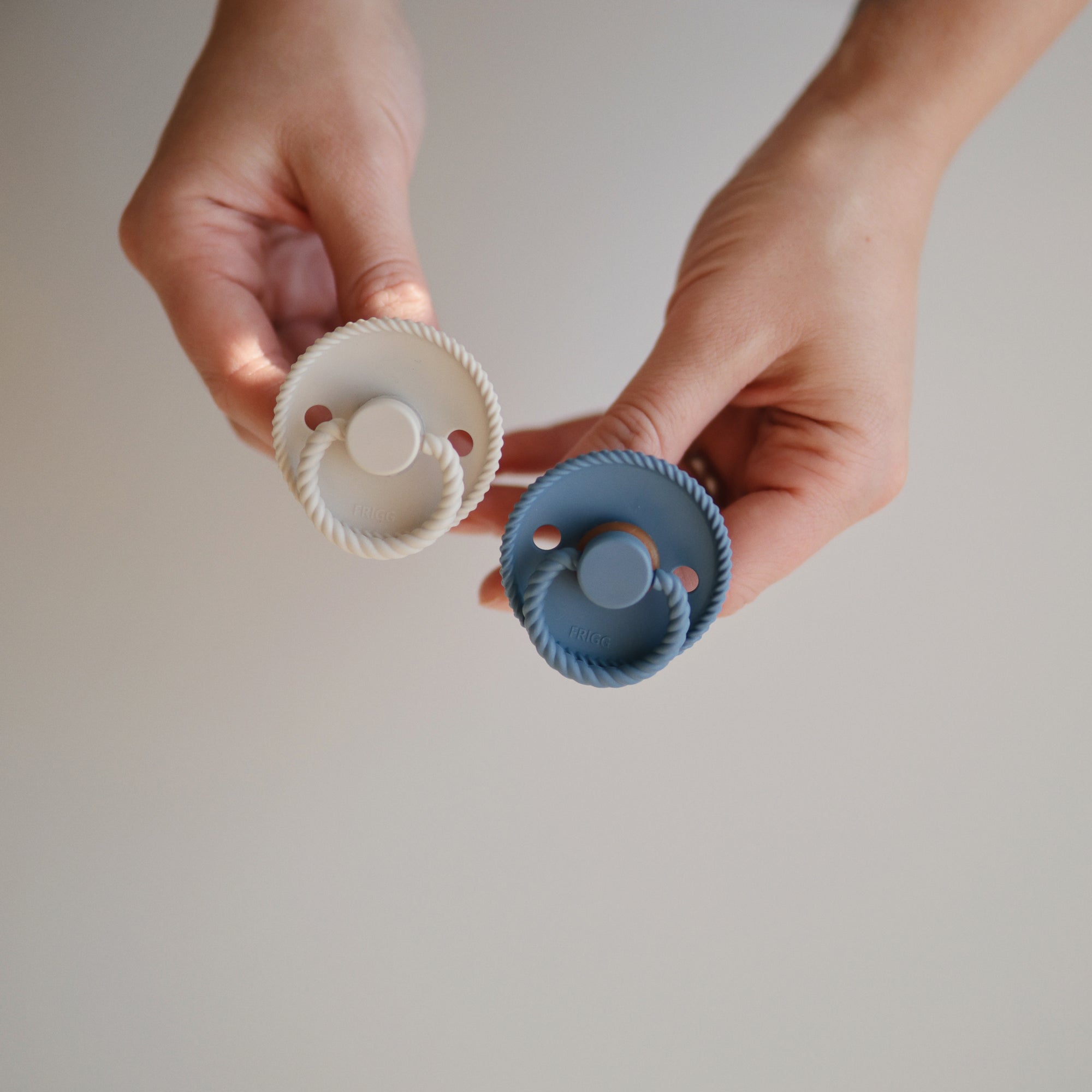 Two hands hold two Mushie FRIGG Rope Natural Rubber Pacifiers—one white, one blue—against a plain background. Each baby essential features a round, textured edge and circular shield with a natural rubber latex nipple. Pack of 2.