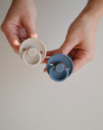 Two hands hold two Mushie FRIGG Rope Natural Rubber Pacifiers—one white, one blue—against a plain background. Each baby essential features a round, textured edge and circular shield with a natural rubber latex nipple. Pack of 2.