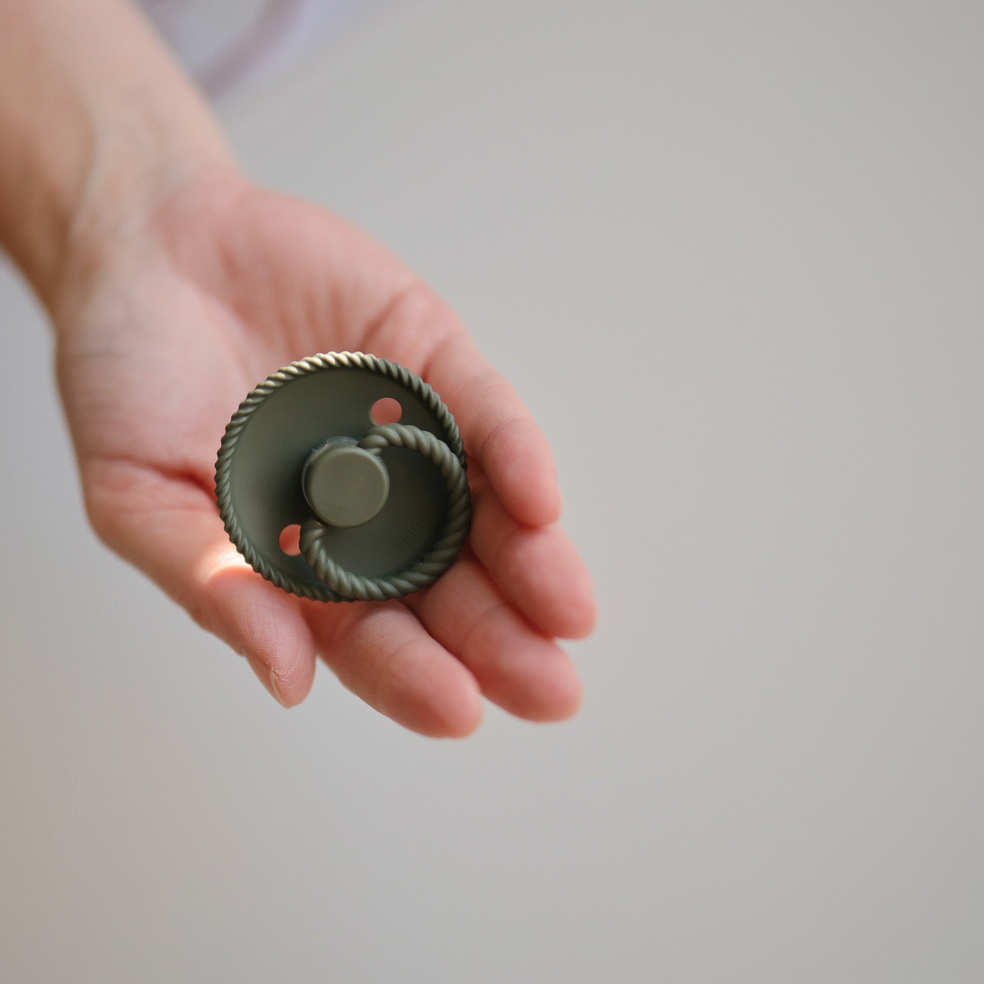A hand holds a FRIGG Rope Silicone Pacifier from the FRIGG Rope Silicone Pacifier 2-Pack, featuring medical grade green silicone with a circular, textured rim and two holes near the center, against a neutral background.
