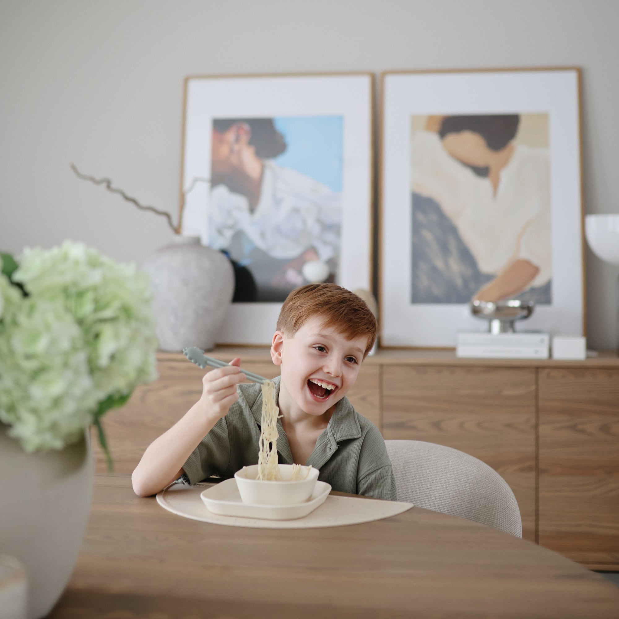 Smiling boy self-feeding with mushie Training Chopsticks at table with art in background.