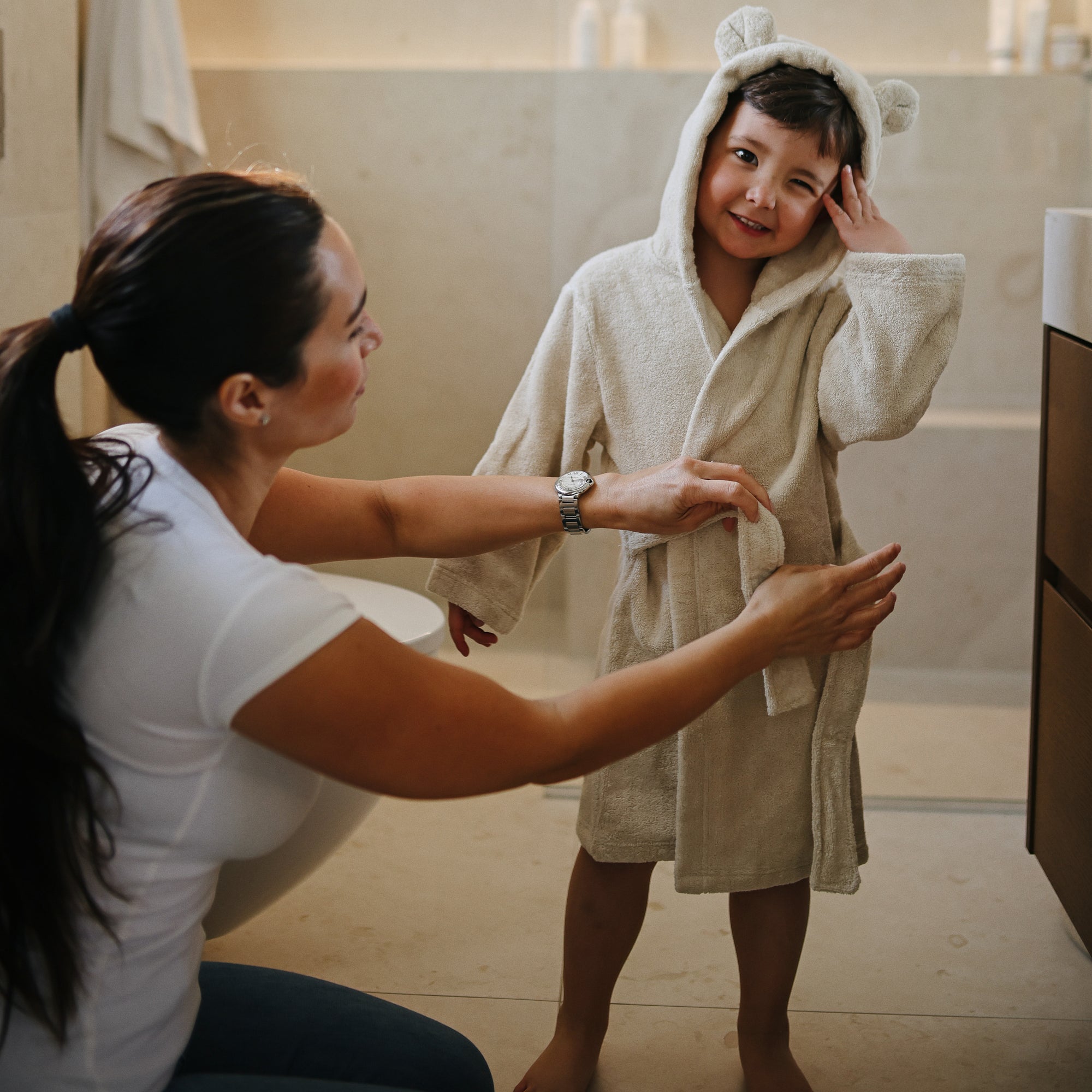A woman helps a smiling child wearing the mushie Bear Robe in a bathroom.