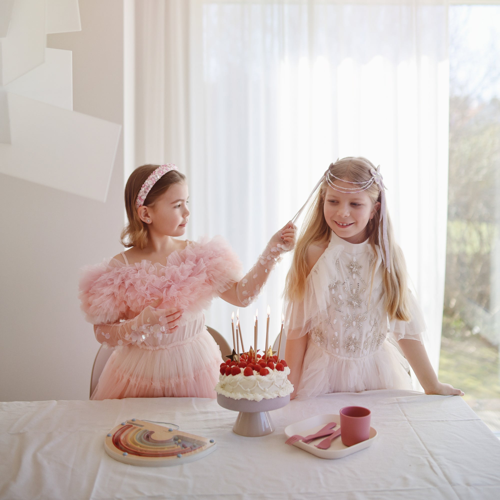 Two girls in pink stand by a birthday cake with a mushie Dinnerware Cutlery Set on the table.