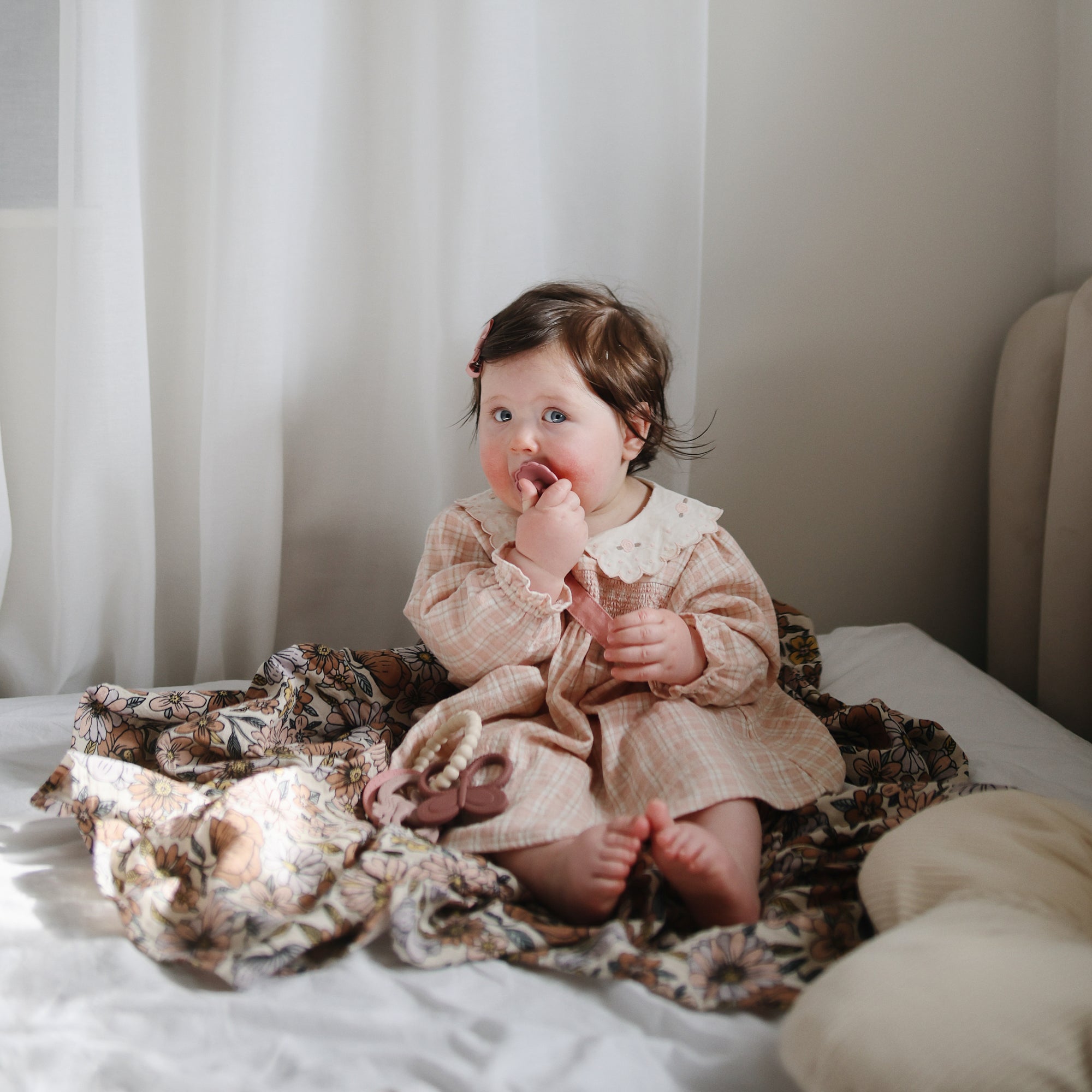 Baby in a pink dress snuggled in a mushie Organic Cotton Muslin Swaddle Blanket on the bed.