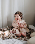 Baby in a pink dress snuggled in a mushie Organic Cotton Muslin Swaddle Blanket on the bed.
