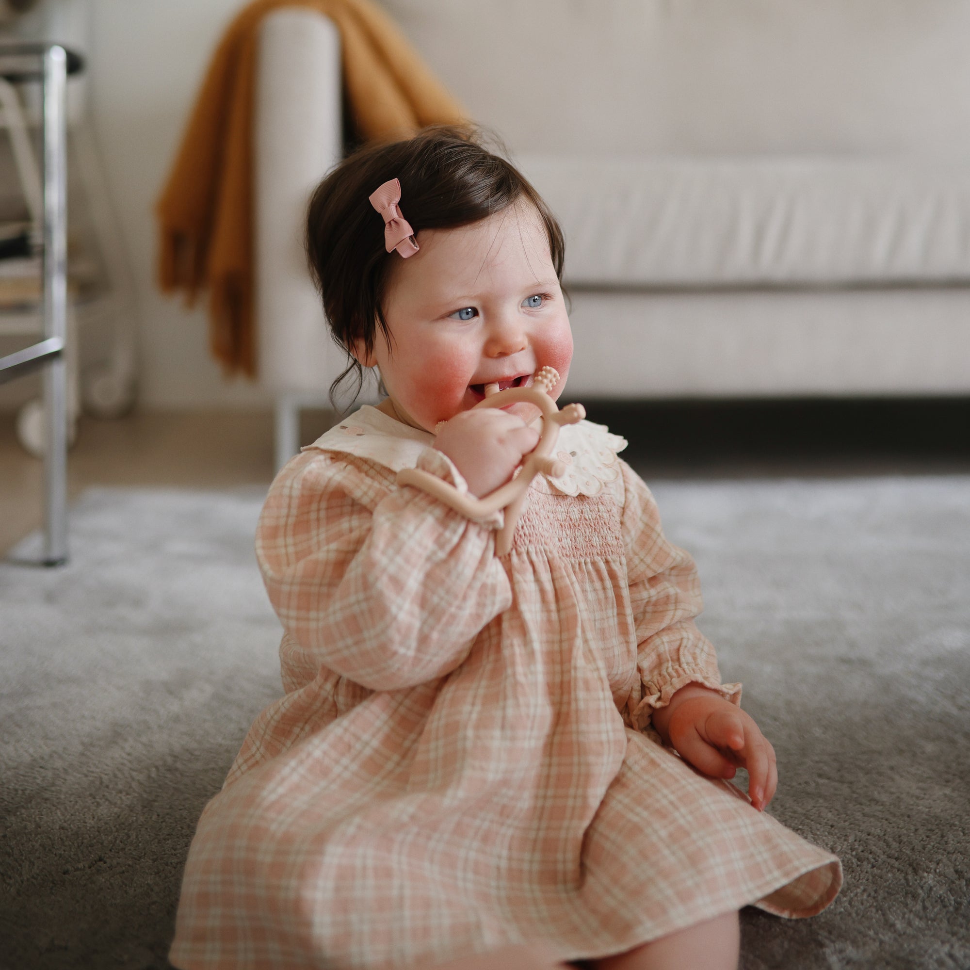 Baby in a plaid dress sits on a carpet, chewing on a mushie No-Drop Sensory Teether.