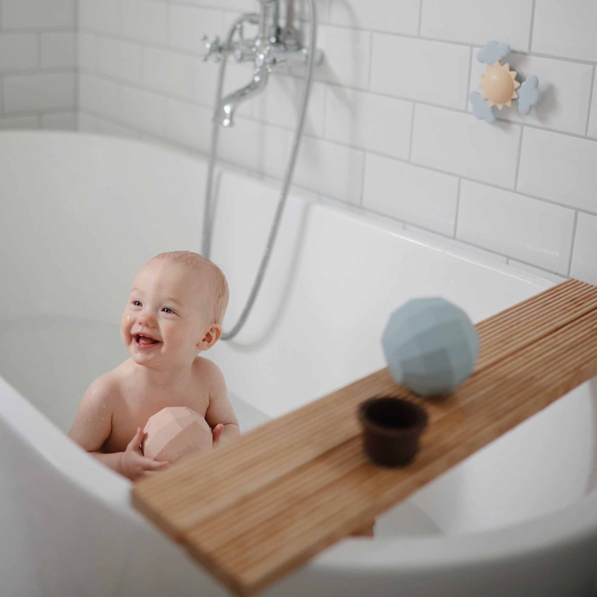 Smiling baby in bath holds mushie Nesting Spheres Sensory Toy; toys on wooden tray nearby.
