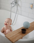 Smiling baby in bath holds mushie Nesting Spheres Sensory Toy; toys on wooden tray nearby.