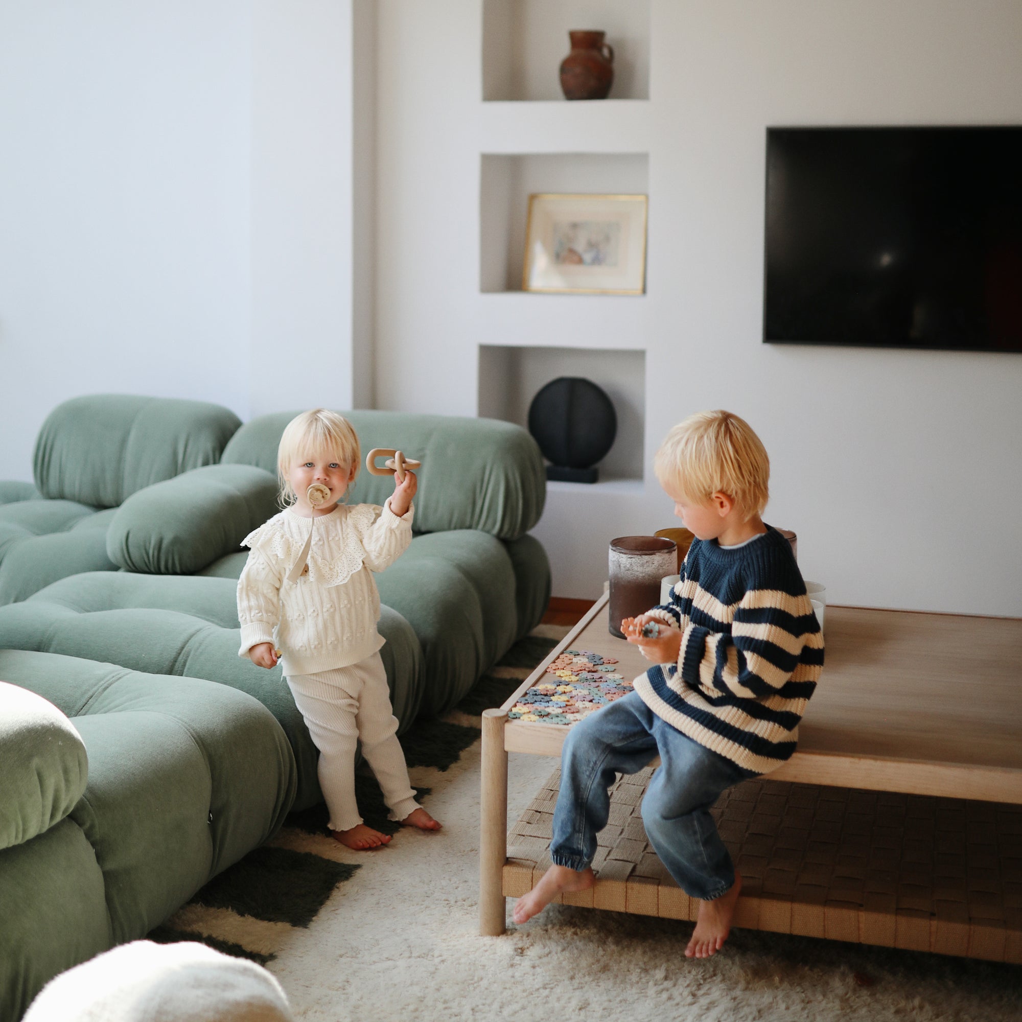 Two children play as a mushie Linen Pacifier Clip 2-Pack sits on the living room table.