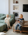 Two children play as a mushie Linen Pacifier Clip 2-Pack sits on the living room table.