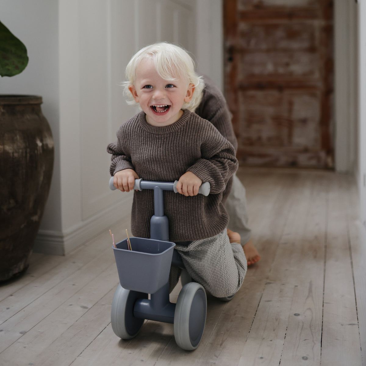 A laughing toddler rides a gray mushie Mini Toddler Bike down a hallway.