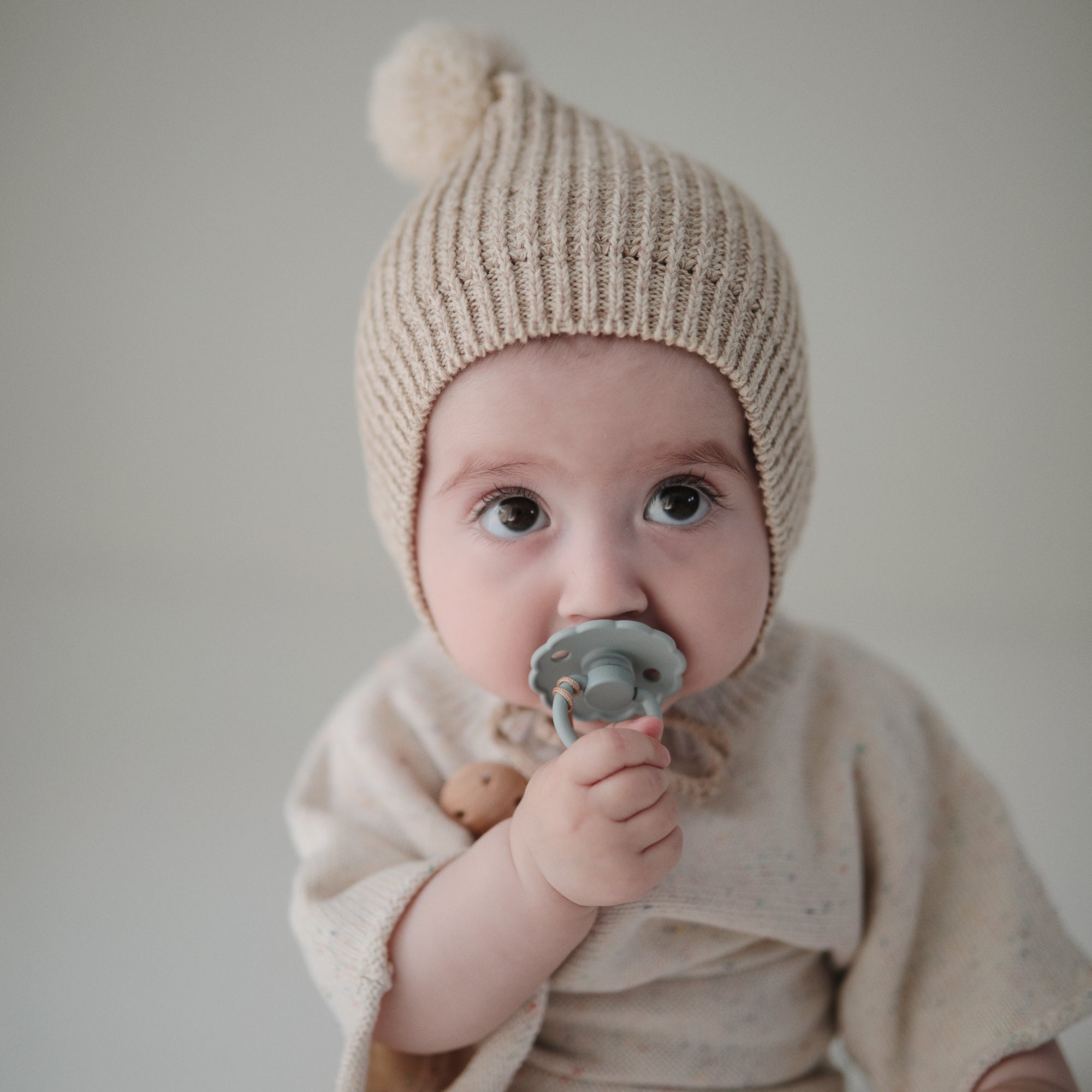 A baby in a beige knit hat and cream outfit holds a gray FRIGG Daisy Silicone Pacifier from the Limited Edition 2-Pack, with a simple, softly blurred background.