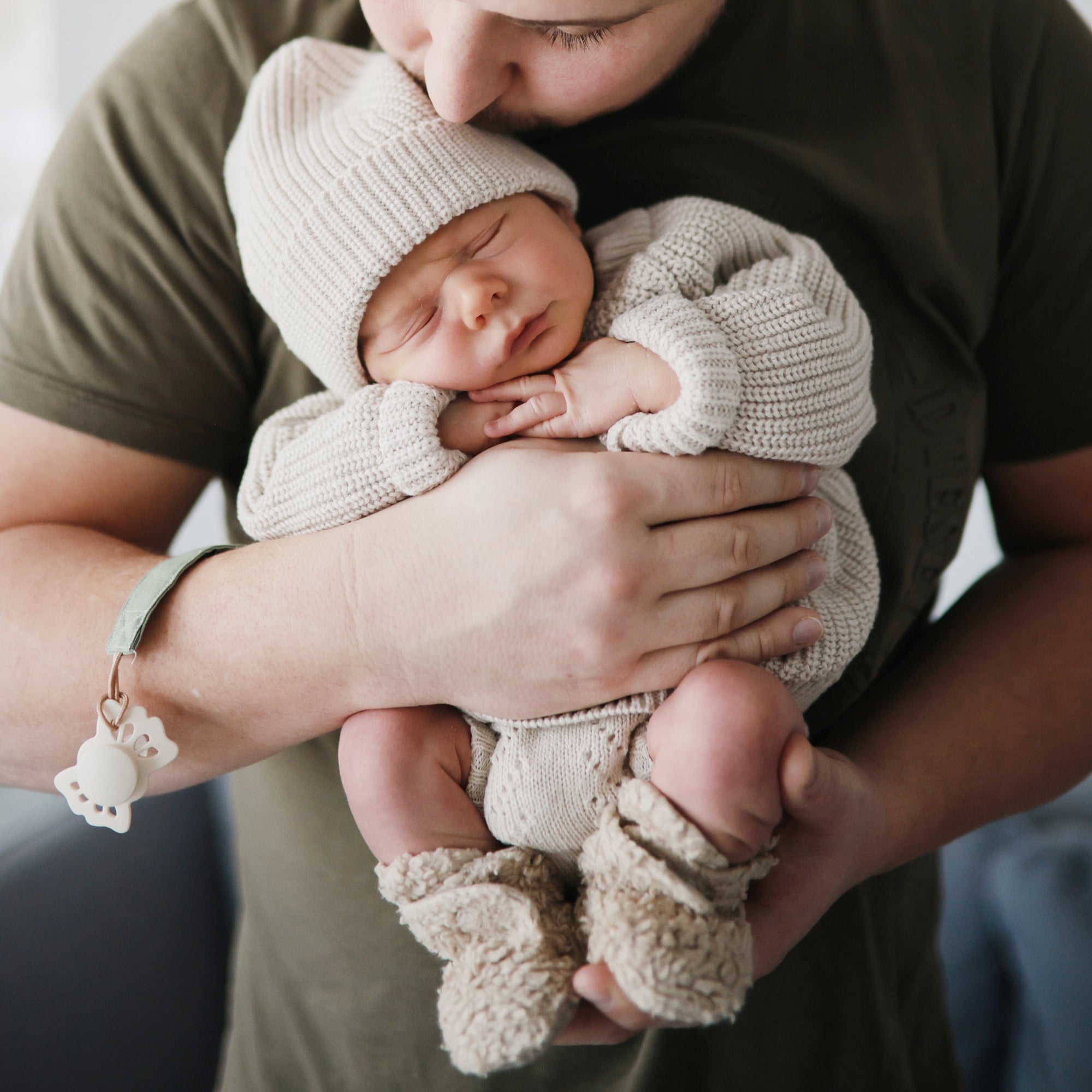 An adult cradles a sleeping baby in knitwear with a mushie Linen Pacifier Clip 2-Pack attached.
