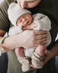An adult cradles a sleeping baby in knitwear with a mushie Linen Pacifier Clip 2-Pack attached.