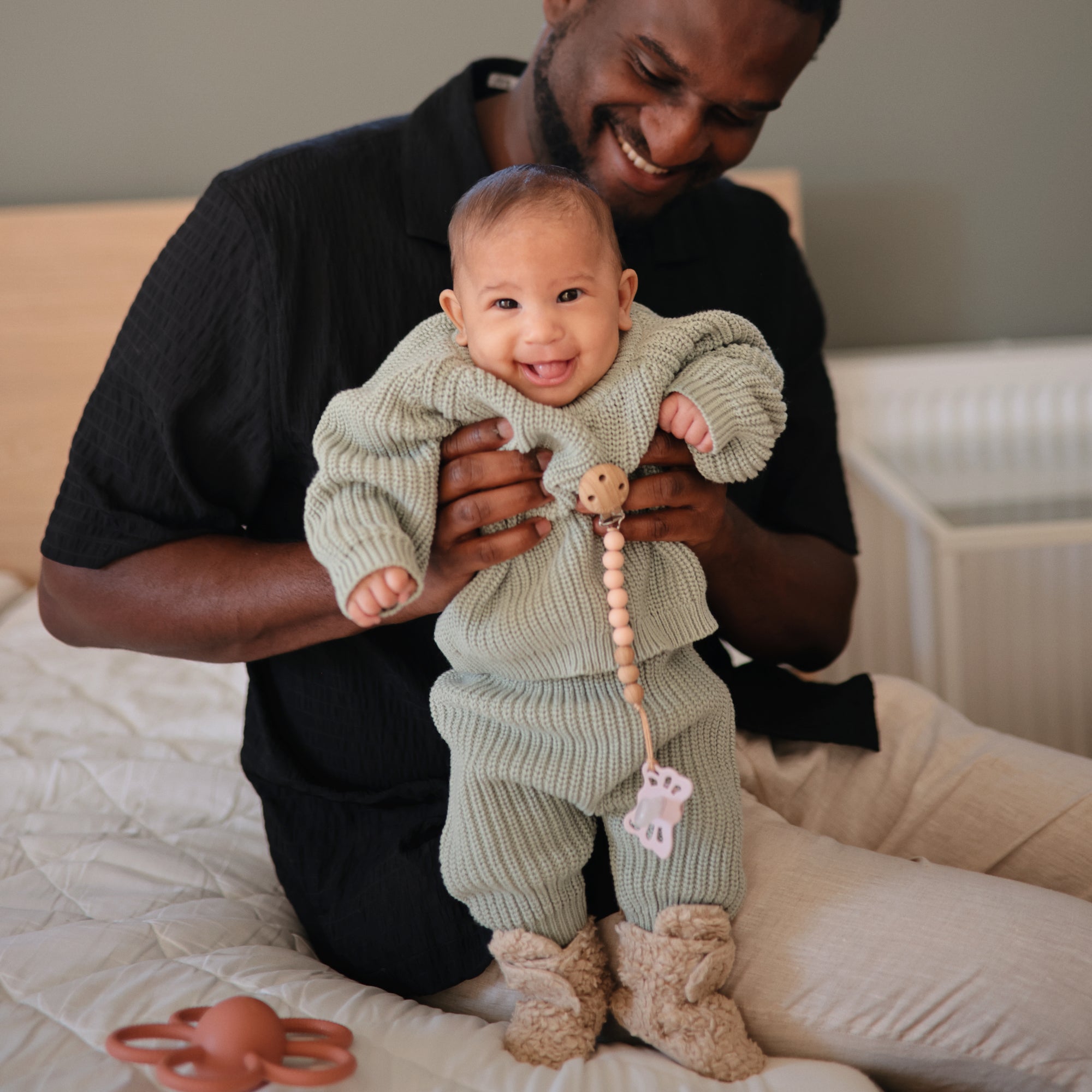 A smiling baby in a green knit outfit and fuzzy boots is held by an adult on the bed. Nearby rests a FRIGG Butterfly Anatomical Silicone Pacifier 2-Pack (0-6 Months) from FRIGG, designed for baby's comfort.
