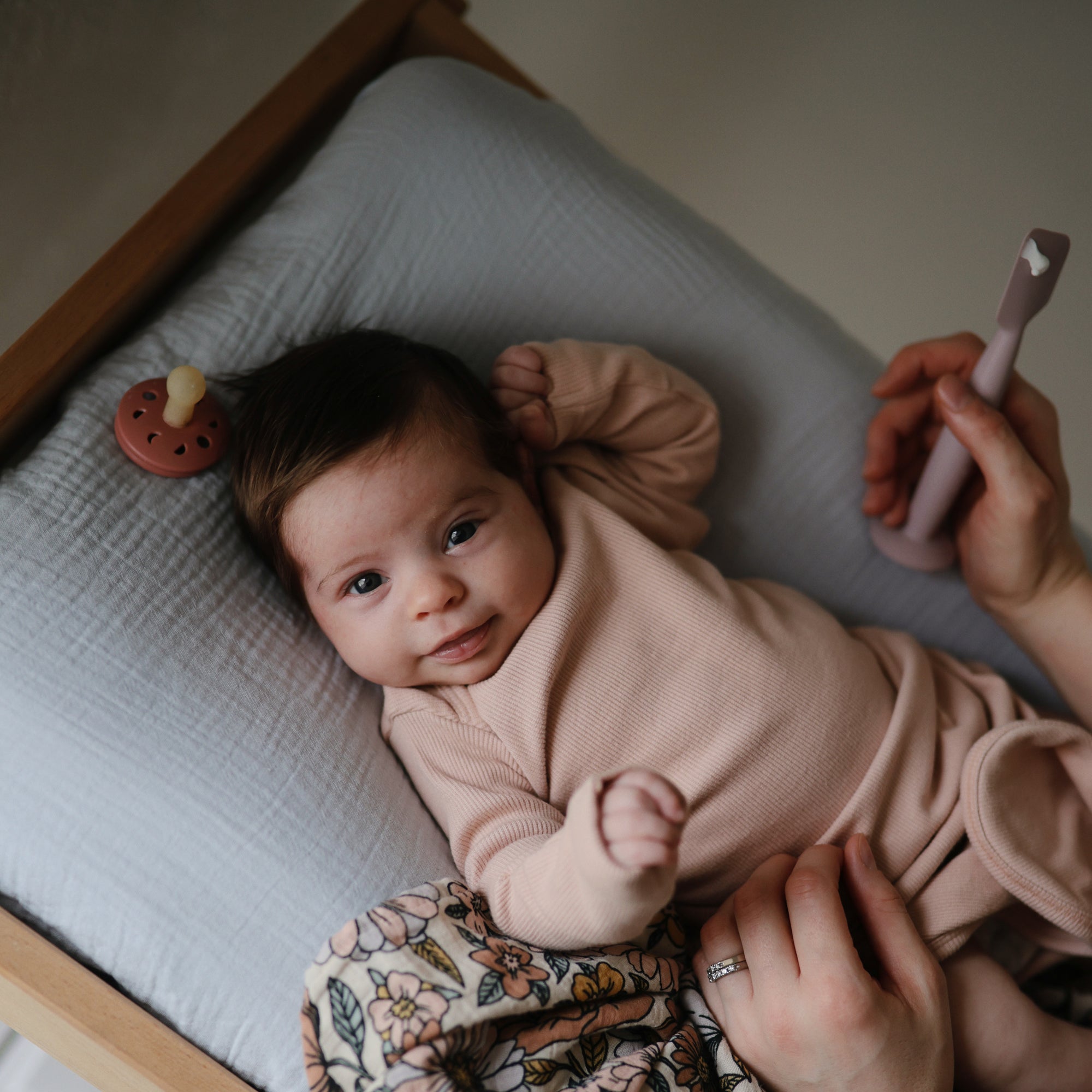 A baby in a light pink outfit lies on a cushioned changing pad, smiling up. An adult hand holds a silicone baby toothbrush, and the FRIGG Moon Natural Rubber Pacifier from FRIGG rests nearby among other baby essentials.