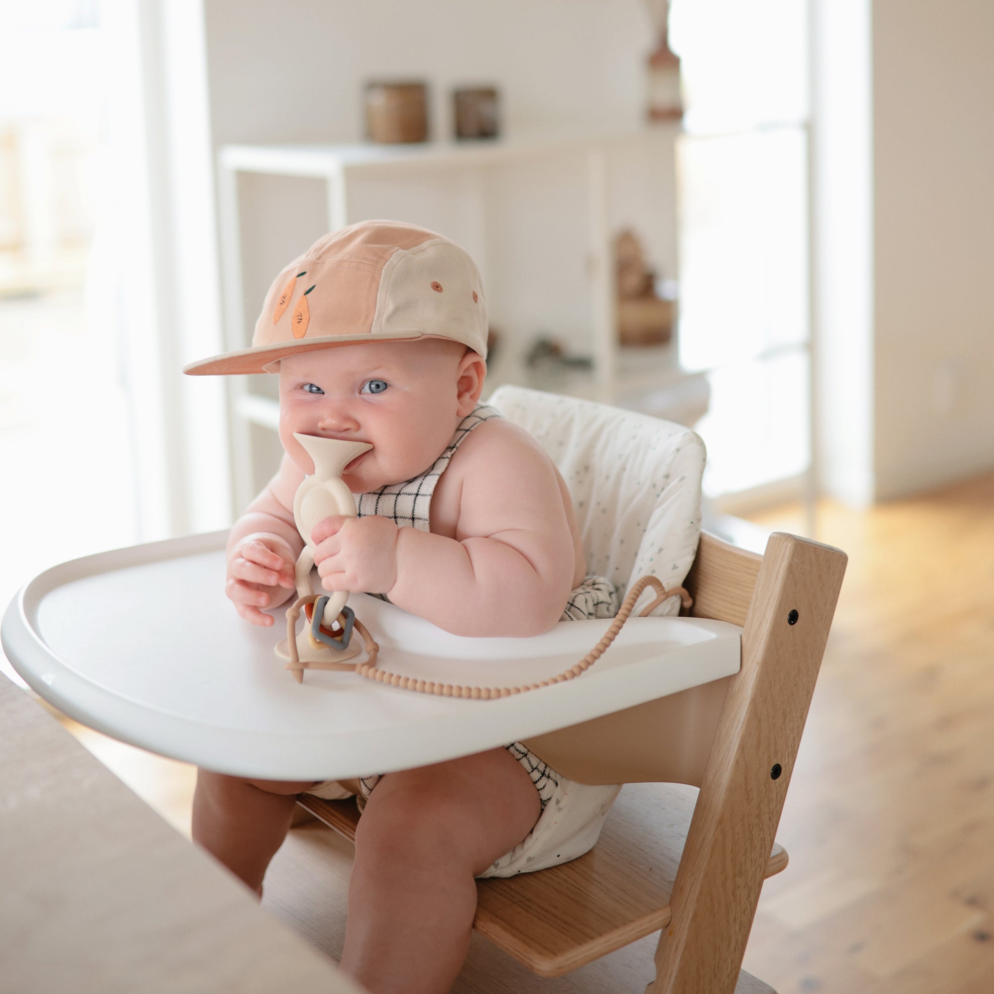 A baby in a high chair, smiling, wearing a cap and holding a mushie HexaPop Suction Toy.