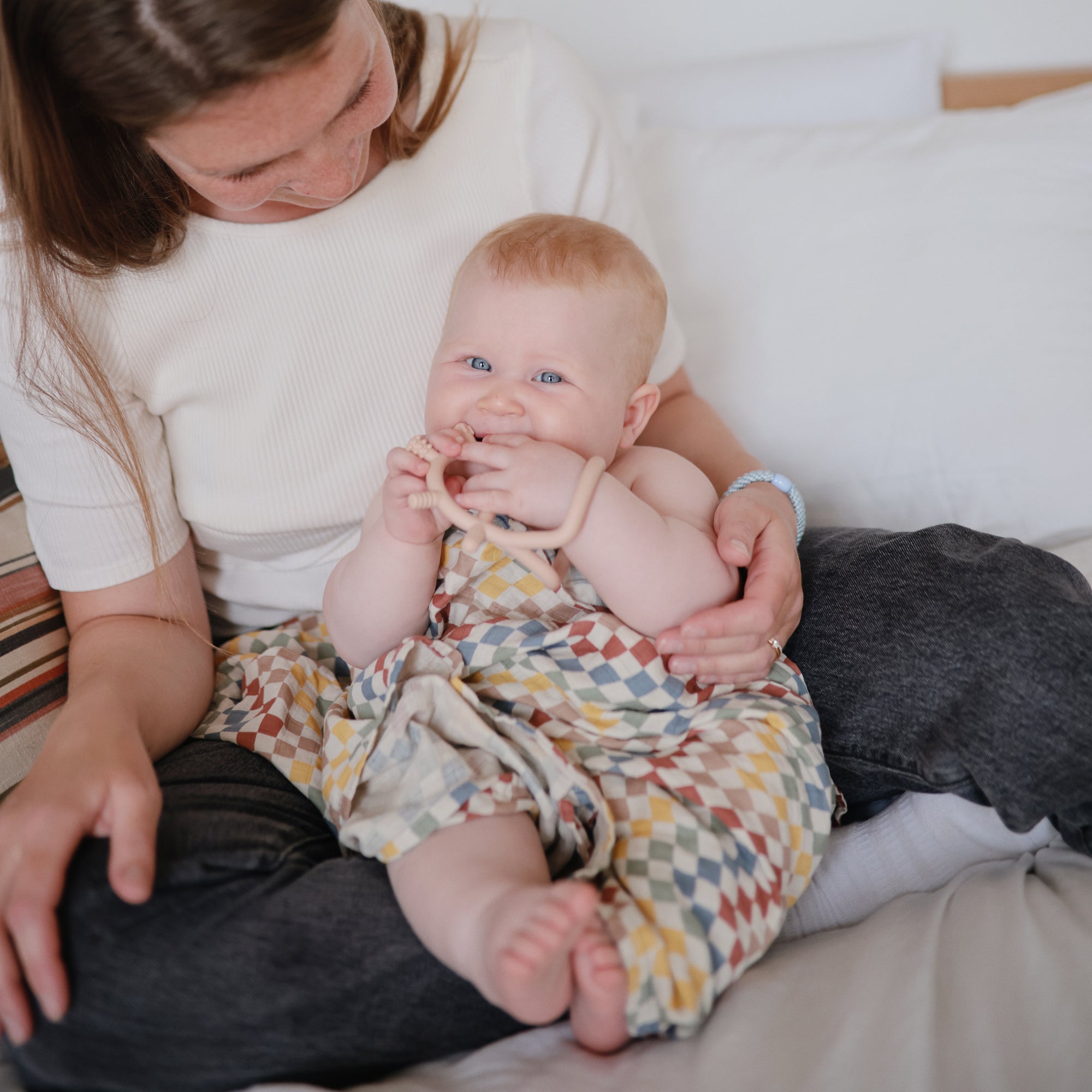 Woman holds smiling baby in romper with mushie No-Drop Sensory Teether.