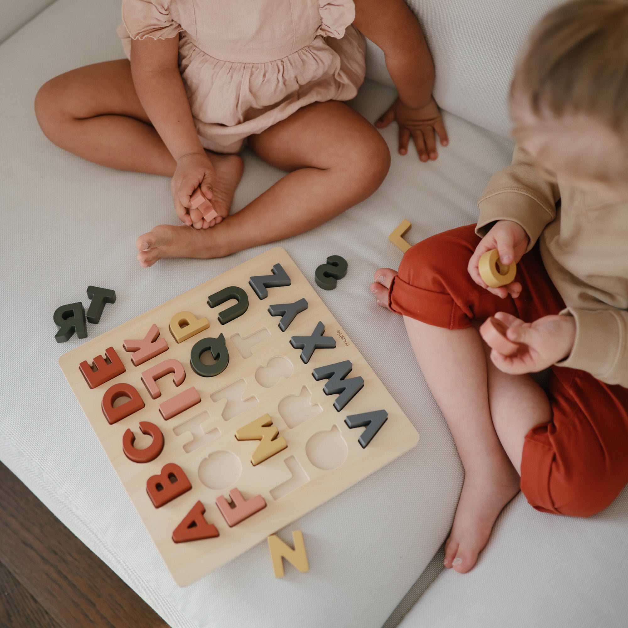 Two young children sit on a white couch playing with a wooden alphabet puzzle. Colorful letters are placed on the board and scattered around them as they interact with the pieces.