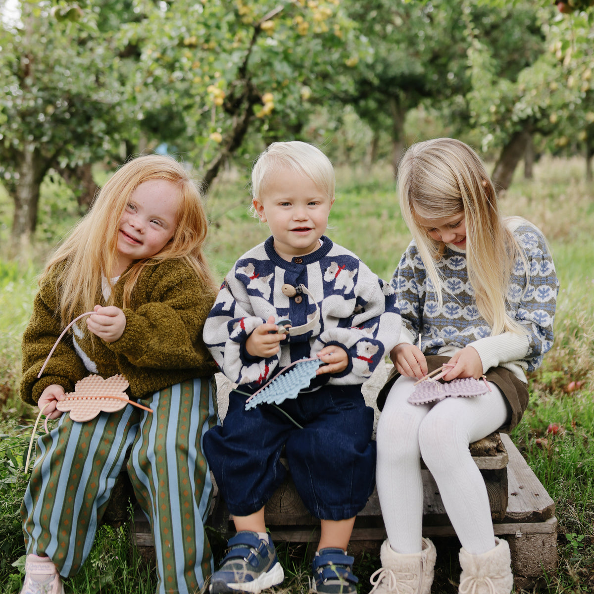 Three young children smile outdoors, playing with mushie Silicone Sensory Toy fine motor crafts.