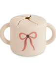 Mushie Snack Cup: Beige, spill-proof, two handles, pink bow design on front.