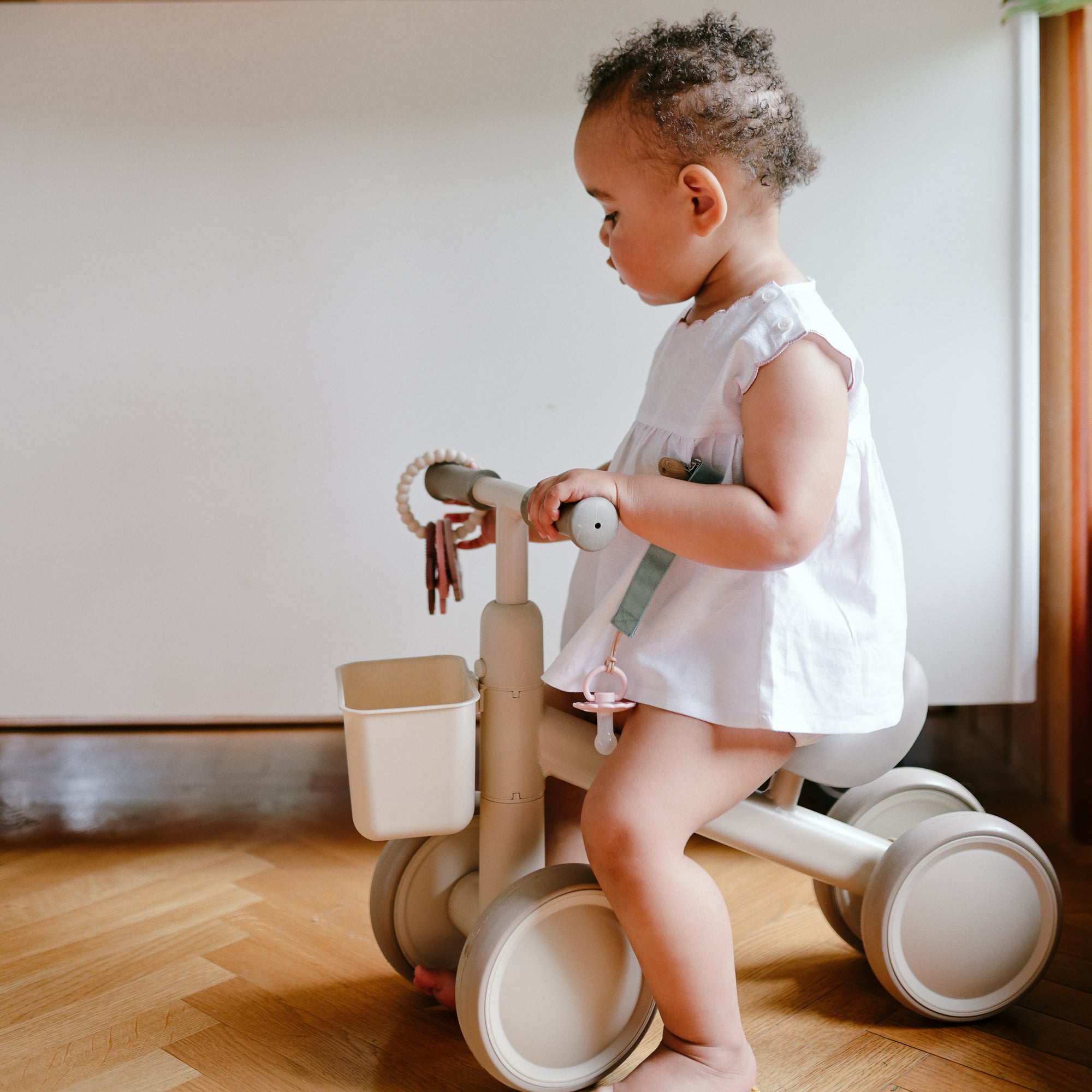 A toddler enjoys her first indoor ride on a beige mushie Mini Toddler Bike in a white dress.