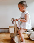 Toddler in white dress holding a mushie Teething Ring, sitting on a beige tricycle indoors.