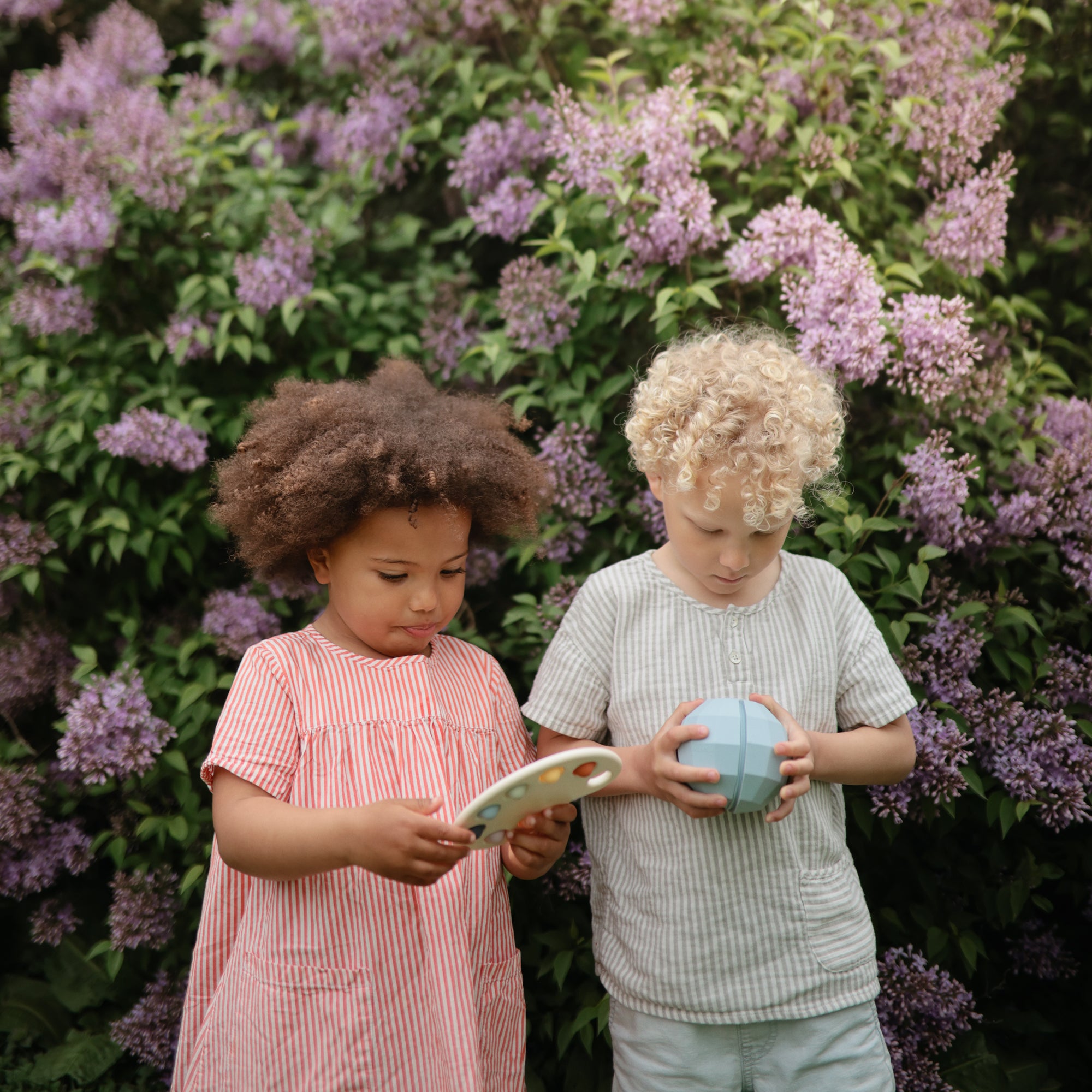 Two children hold mushie Nesting Spheres Sensory Toys, wearing striped summer outfits.