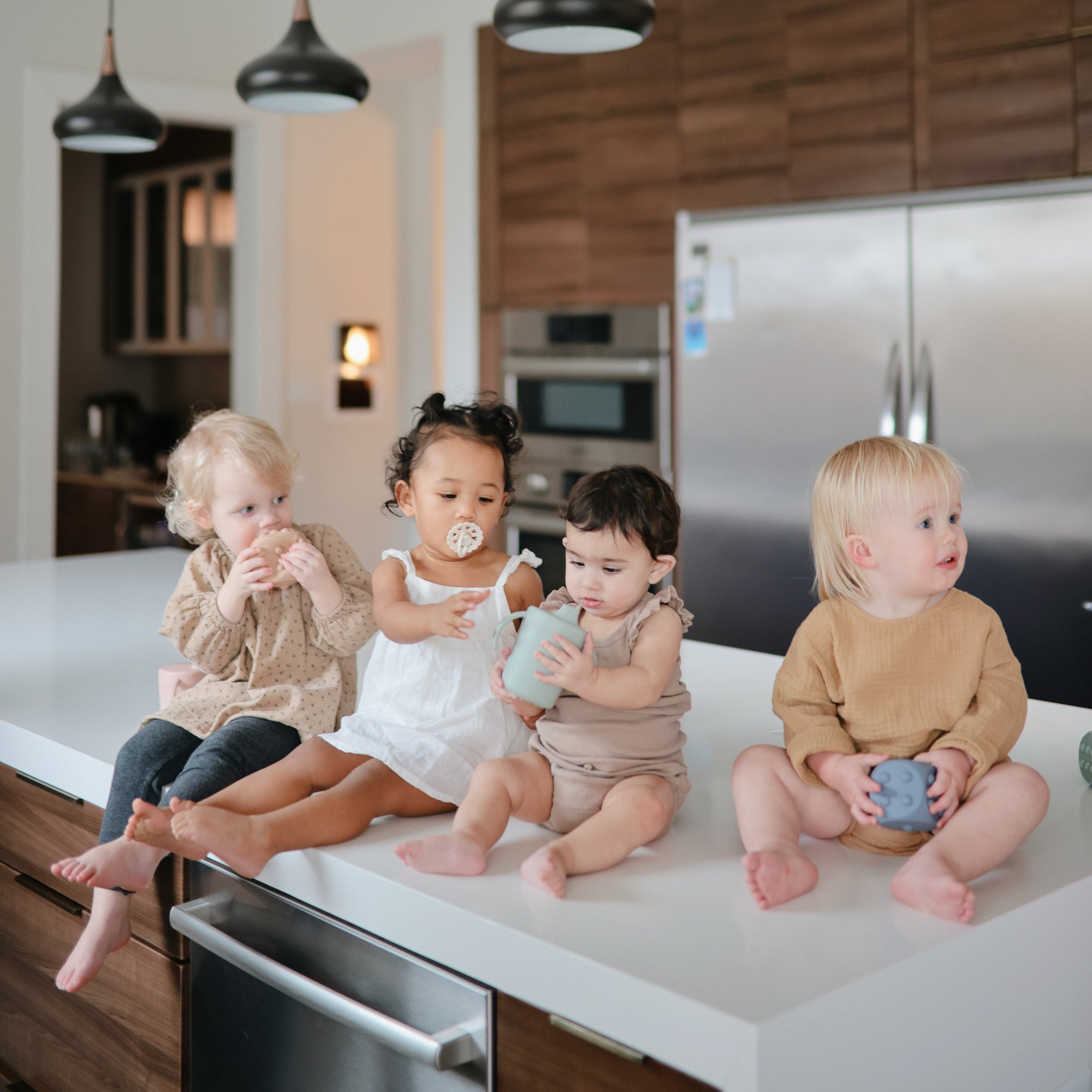 Four toddlers sit on a kitchen island with cups, cookies, and pacifiers. Nearby, FRIGG Andersen Fairytale Silicone Pacifier 2-Pack by FRIGG—made in Denmark—rests on the counter amid sleek appliances and wooden cabinets.