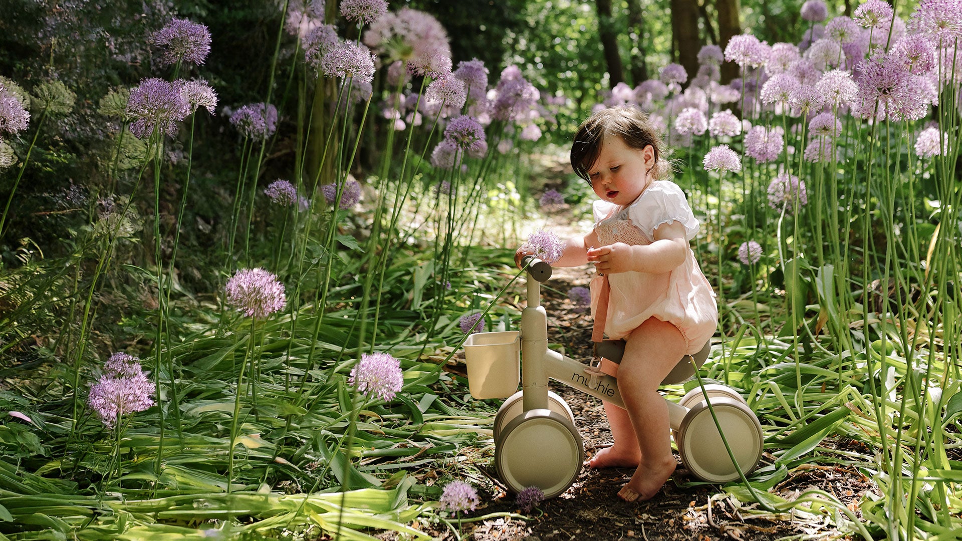 Toddler riding a beige Mushie Mini Toddler Bike on an outdoor path surrounded by purple flowers and greenery.