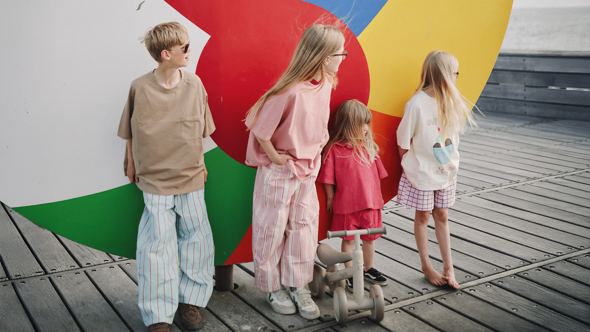 Four children standing on a pier looking out at the water, with a Mushie toddler bike placed between them.
