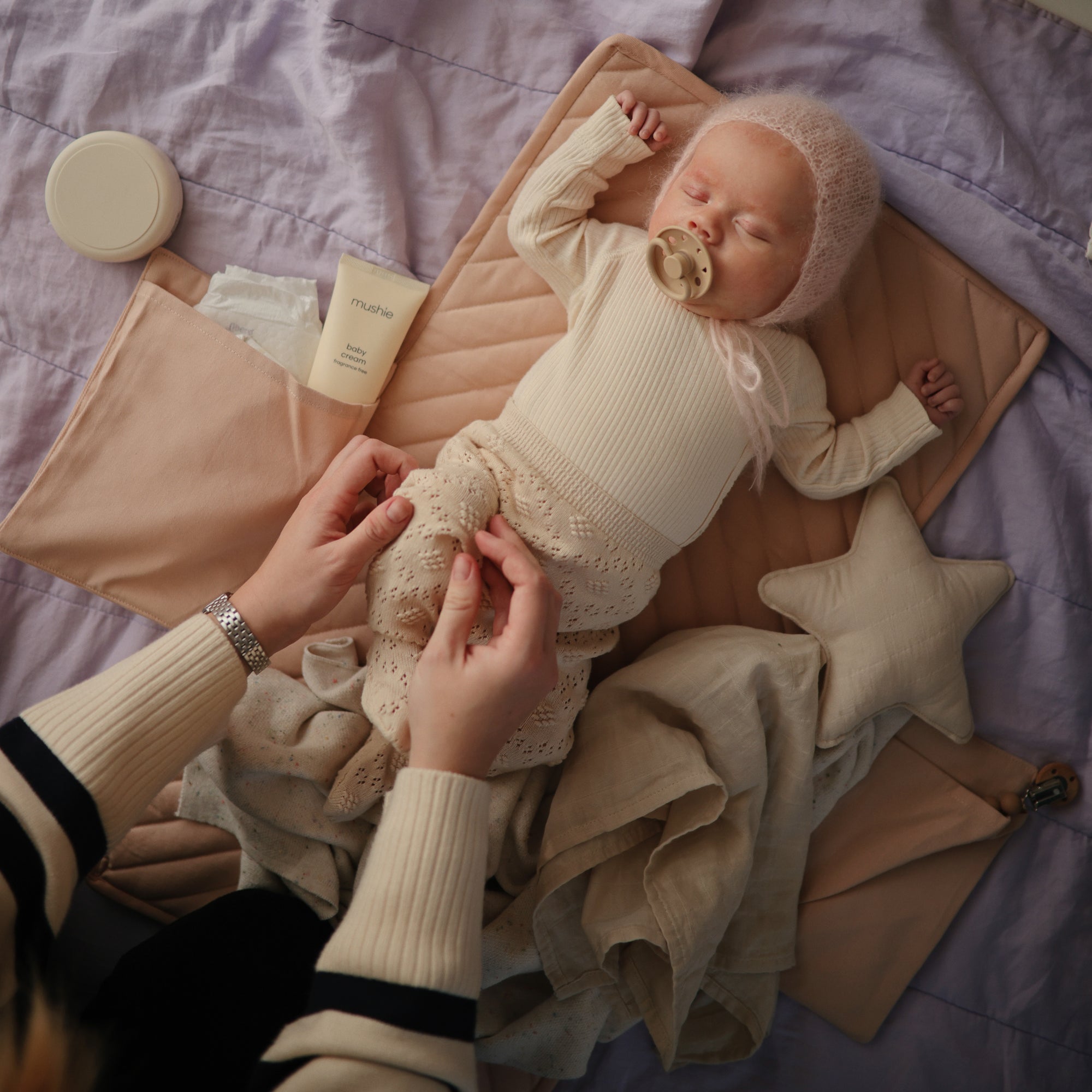An adult applies mushie Baby Cream to a baby with dry skin, who is holding a pacifier.
