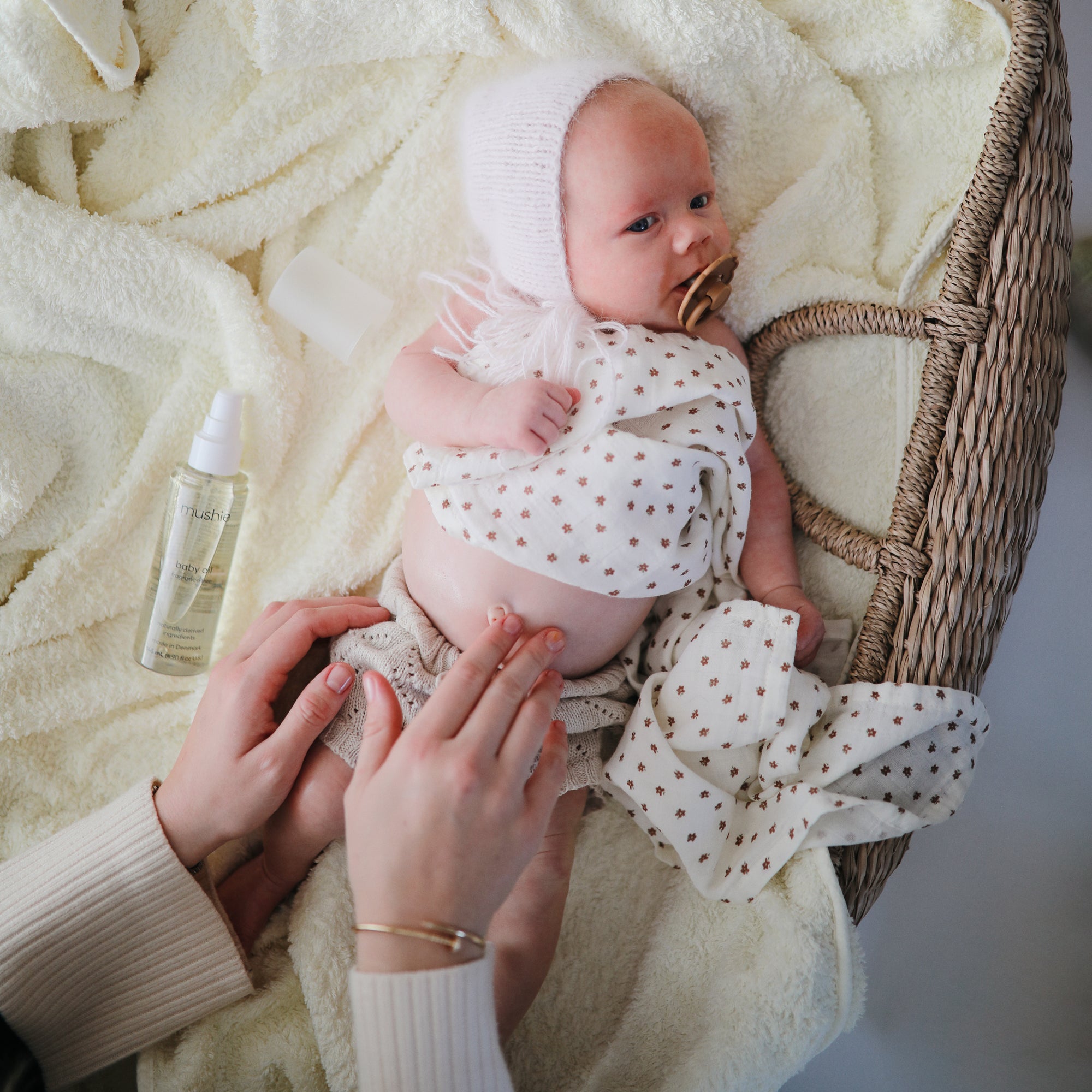 Baby in a basket as someone applies mushie Baby Oil to their belly, wearing a hat and pacifier.