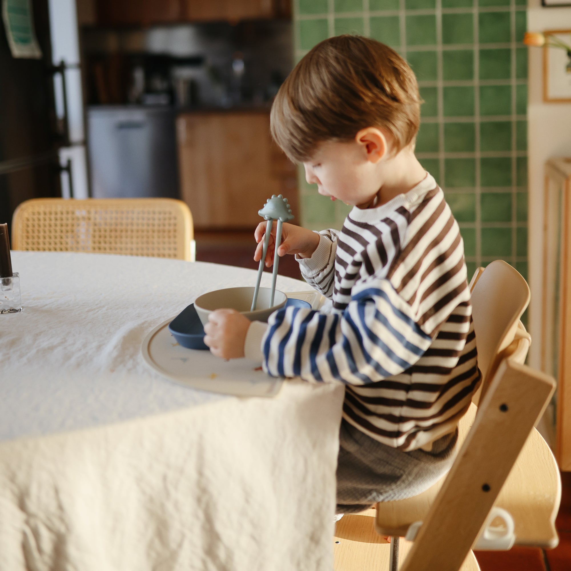 Child eating with chopsticks from mushie Round Dinnerware Bowl at the table.
