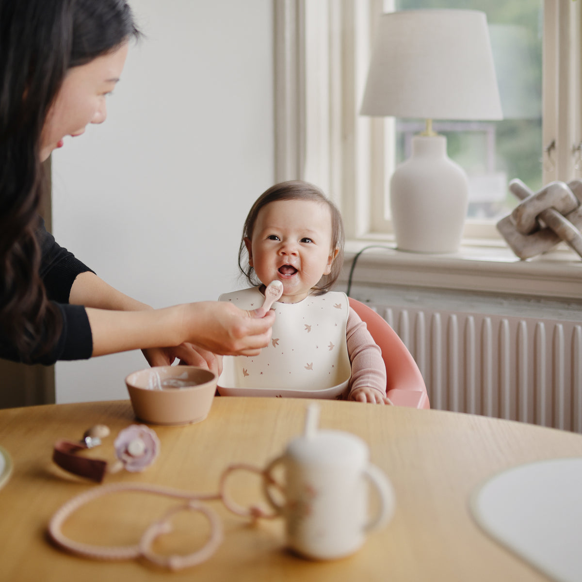 Woman feeds smiling teething baby in high chair with Mushie First Feeding Baby Spoons by window.